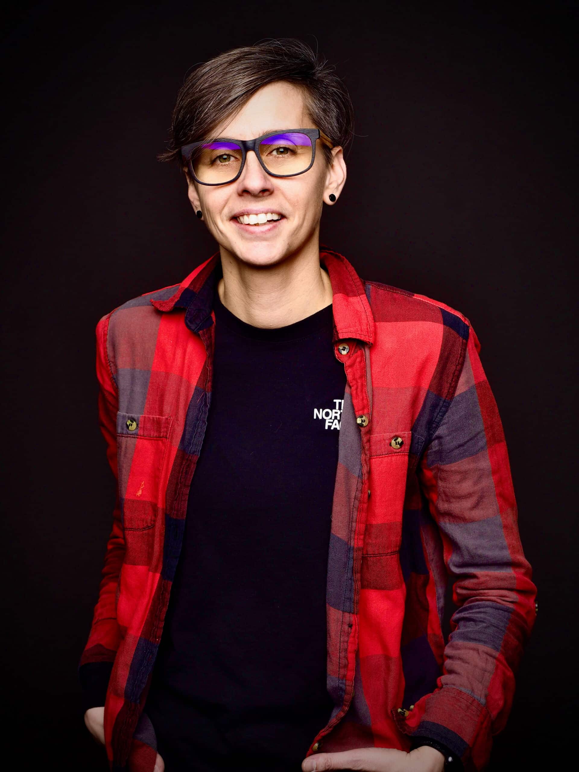Personne aux cheveux bruns courts, portant des lunettes et une chemise à carreaux rouges par-dessus un t-shirt noir, souriante sur un fond sombre, les mains dans les poches.